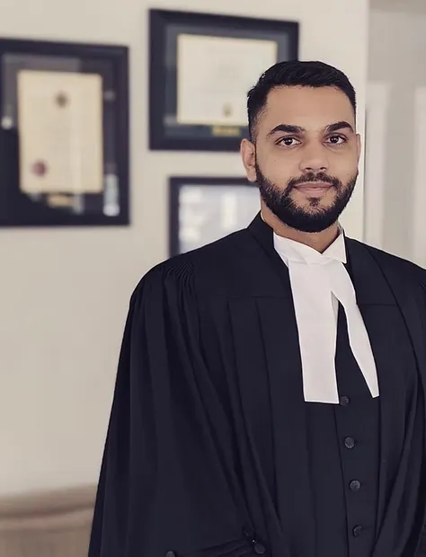 A person wearing legal robes stands indoors in front of framed certificates on a wall.