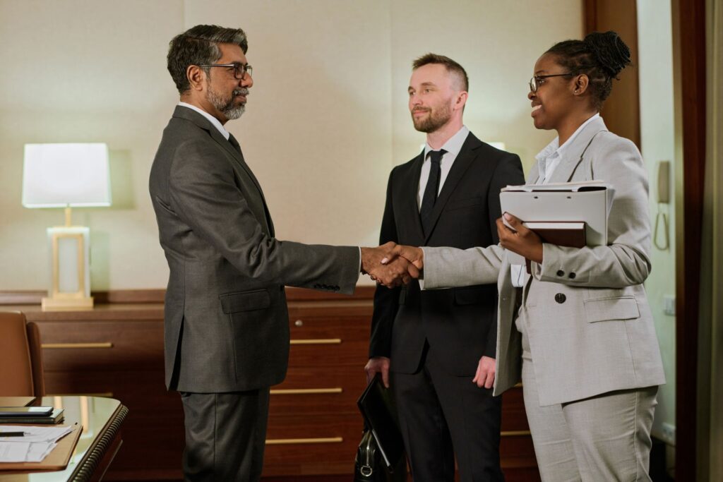 Three business professionals in suits meet in an office. Two are shaking hands while the third stands nearby holding a briefcase, observing the interaction.
