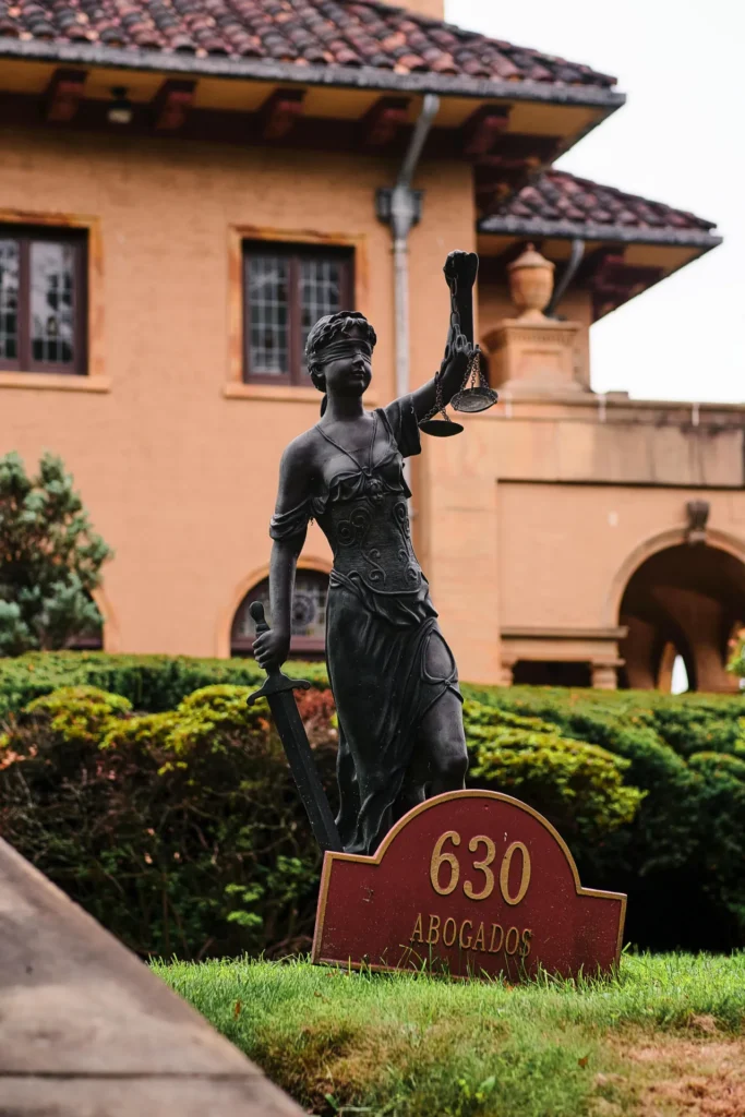 A statue of Lady Justice holding scales and a sword stands in front of a building, with a sign reading "630 Abogados" at its base.