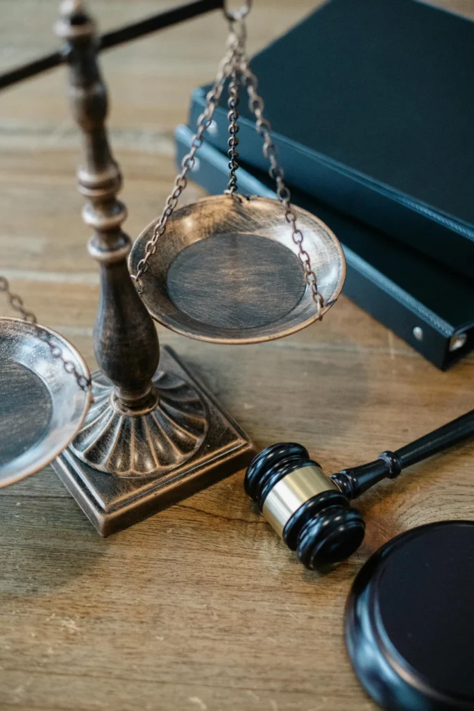 A balance scale, two black binders, and a judge's gavel rest on a wooden table.