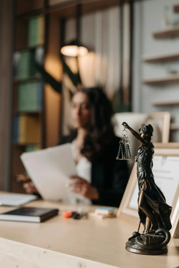 A bronze Lady Justice statue is on a desk with papers and books, while a person in business attire sits in the background holding documents.