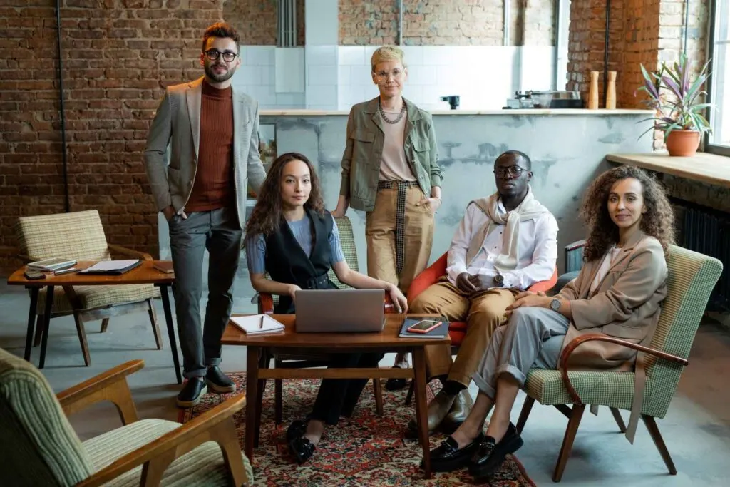 Five people in business attire sit and stand around a coffee table with a laptop, discussing how hiring a family lawyer can help save money, all in a modern office with brick walls and retro furniture.